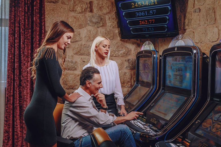 Две красотки, шатенка и блондинка, наблюдают как зрелый привлекательный мужчина играет на автомате в казино