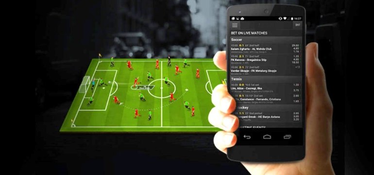 Рука держит смартфона, на экране которого открыт сайт букмекерской конторы, а на заднем фоне виртуальное футбольное поле