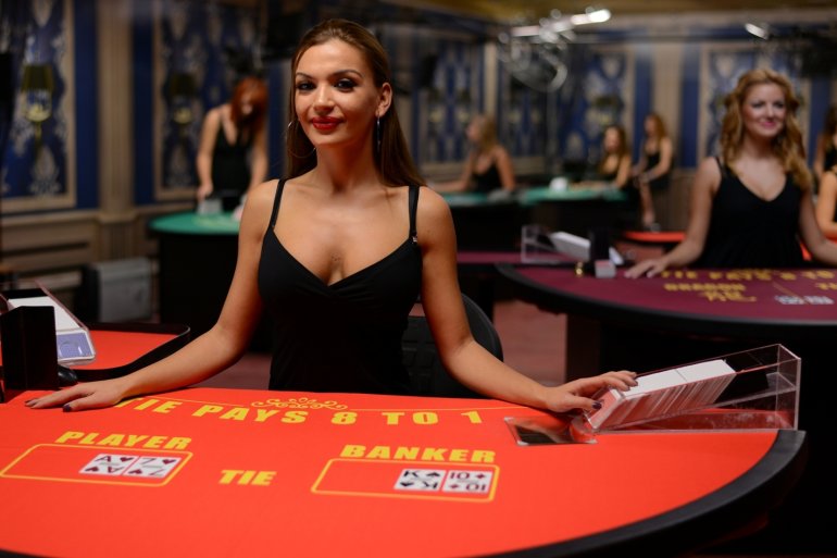 Грудастая девушка крупье в обтягивающем черном платье сидит за столом для баккара в ожидании трансляции игры